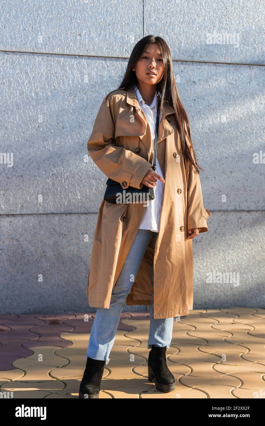 Full body of serious Asian female in stylish outfit with long hair looking at camera while standing near modern building Stock Photo