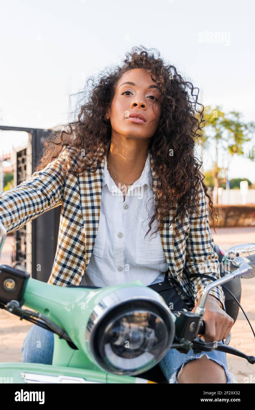 Young wistful black female in trendy outfit looking away while riding scooter on sunny day Stock Photo