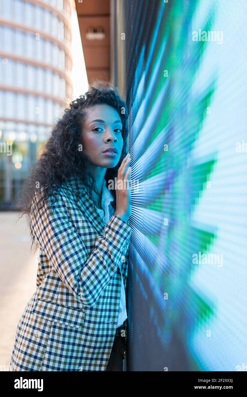 Wistful black female in casual clothes with curly hair looking at camera while leaning on wall with neon illumination Stock Photo