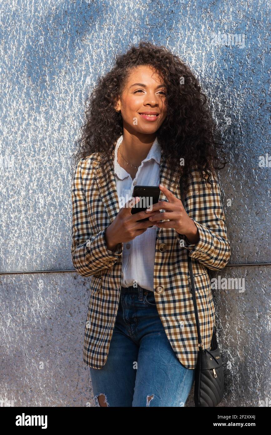 African American female in trendy outfit with curly hair browsing phone while standing on street near concrete wall Stock Photo