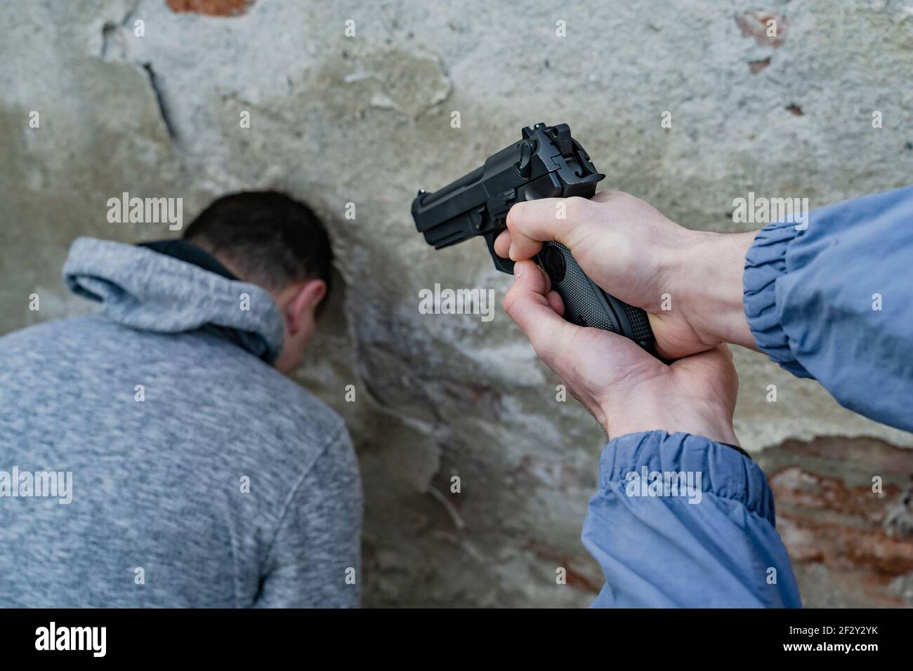 Close up on hand of unknown man terrorist or murderer holding a gun pointed to the head of unknown victim hostage with head leaned against a wall - te Stock Photo
