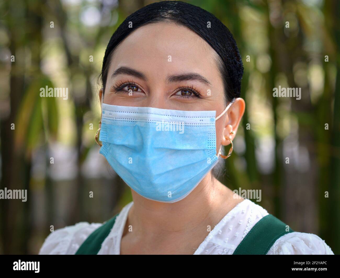 Young Mexican woman smiles with her beautiful brown eyes and wears a light-blue disposable surgical face mask during the coronavirus pandemic. Stock Photo