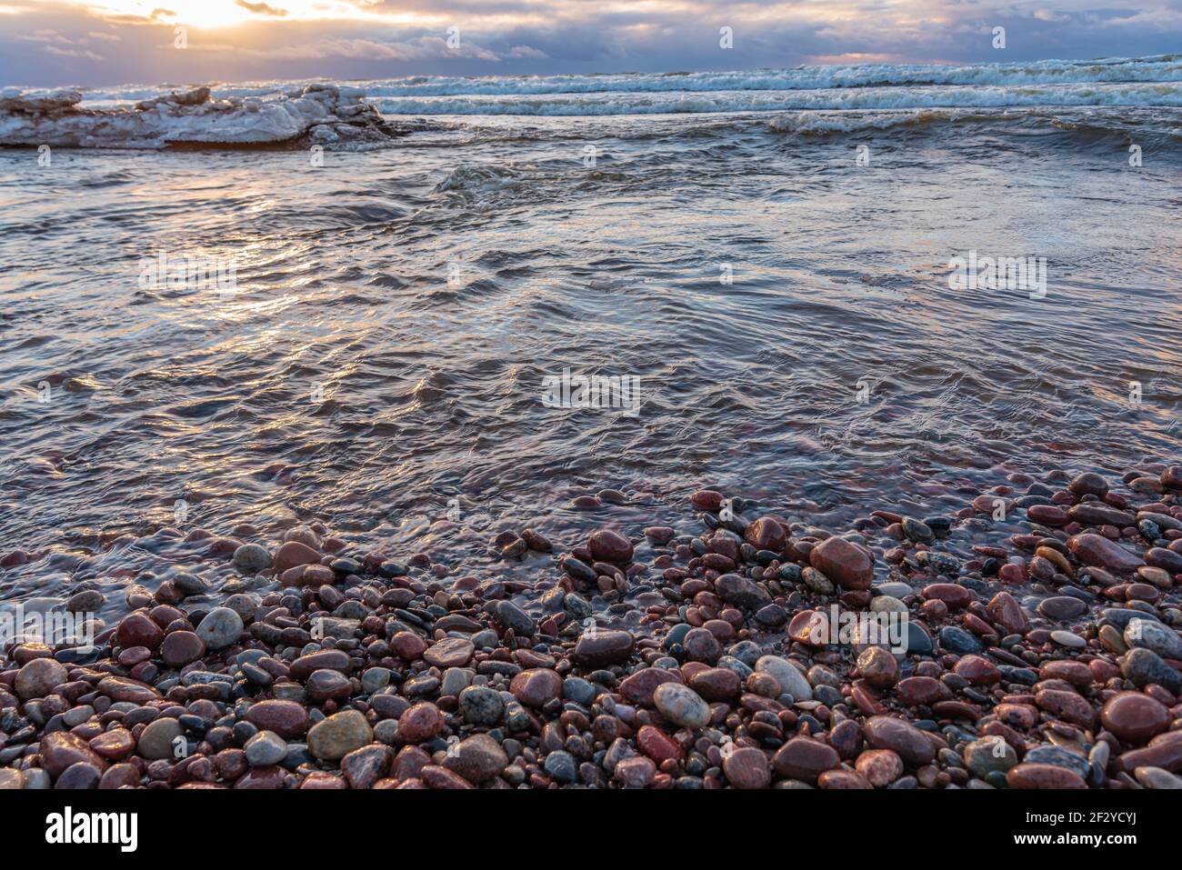small stones on the sea shore with a sunset that illuminates the surface of the water and illuminates the colored stones and clouds in the sky Stock Photo