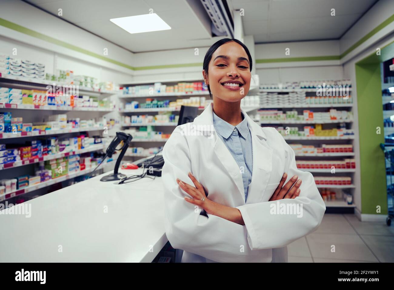 Proud young female with folded hands in labcoat standing in chemist near counter Stock Photo