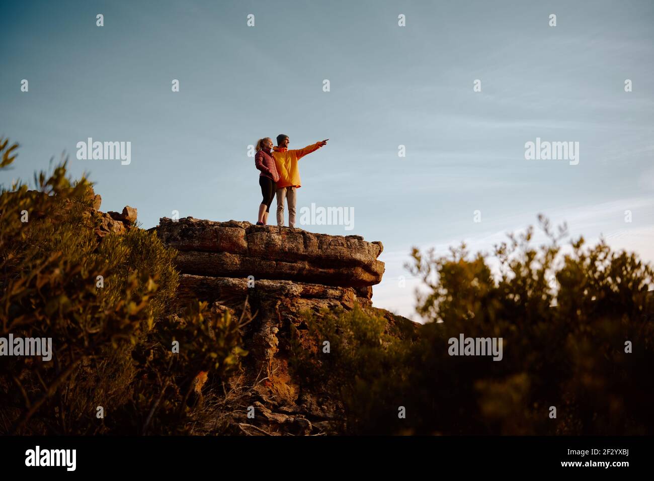 Young man standing at cliff peak pointing and showing something to woman during sunrise Stock Photo