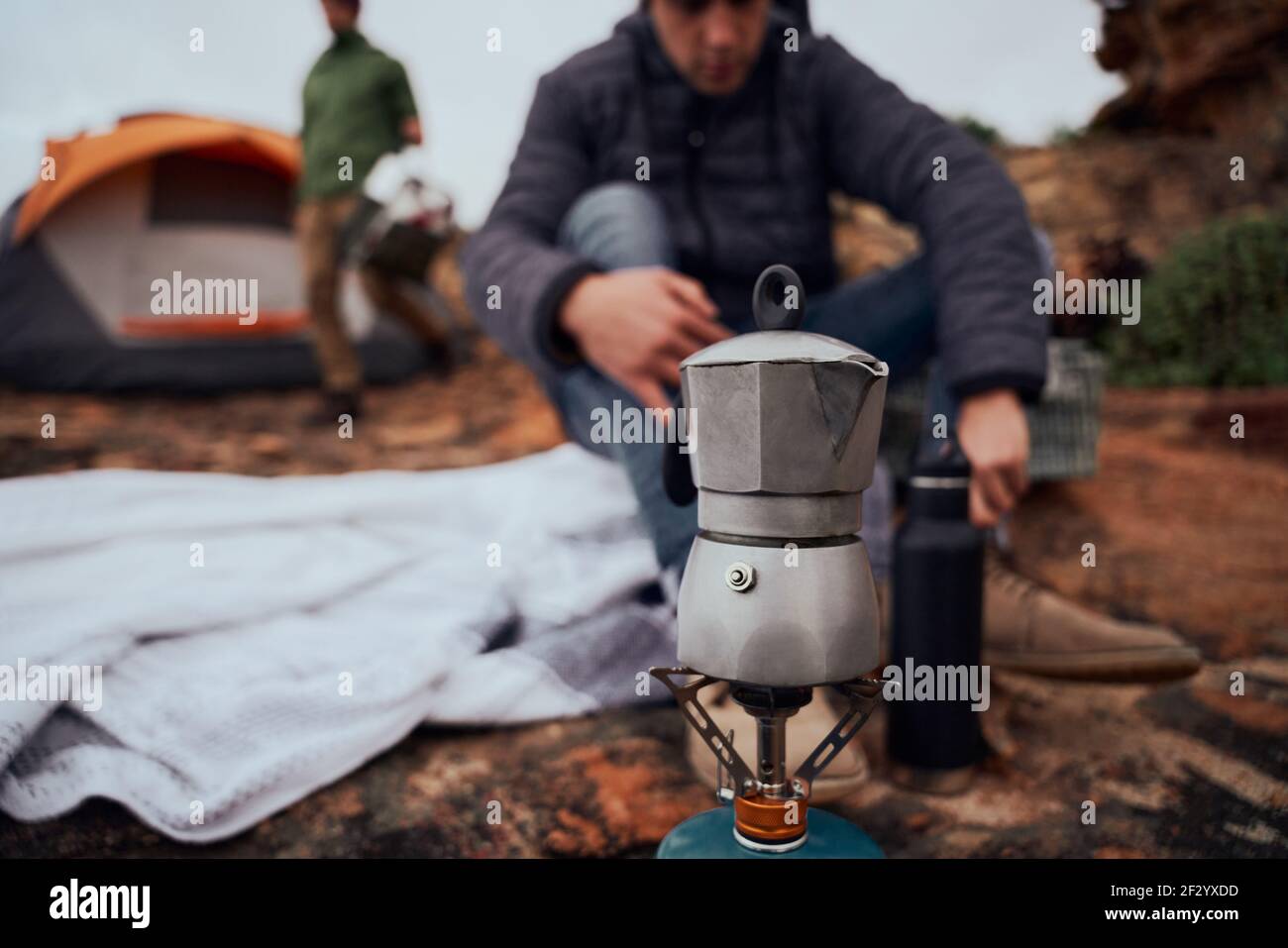 Closeup of kettle on stove during camping while man preparing morning drink sitting on blanket outside tent Stock Photo