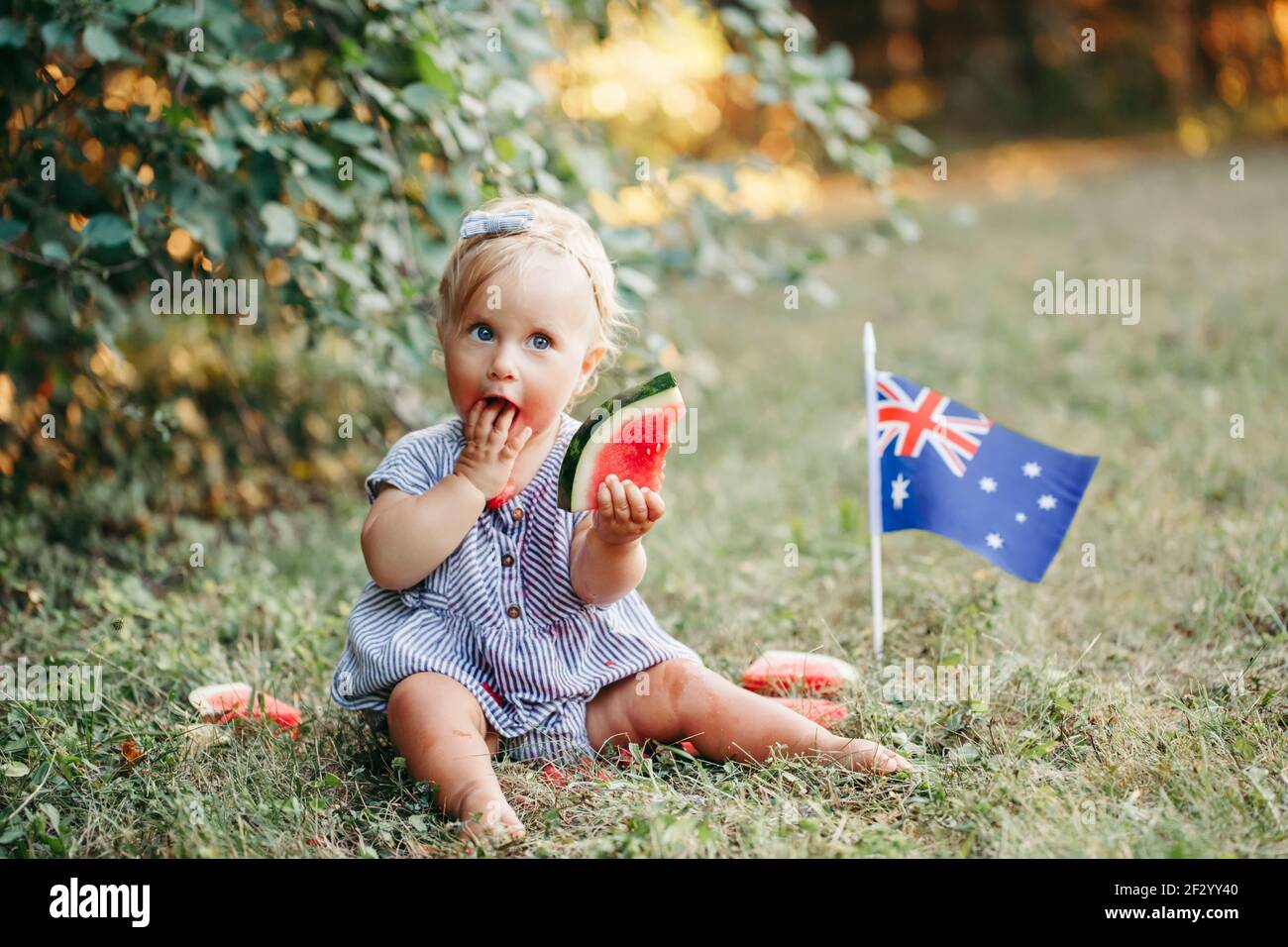 Cute adorable Caucasian baby girl eating ripe watermelon with Australian flag on ground. Funny child kid sitting in park with fresh fruit celebrating Stock Photo