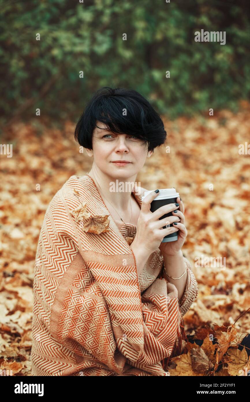 Middle age Caucasian brunette woman with short bob hair wrapped in woollen scarf outdoor. Woman sitting among autumn fall yellow leaves in park Stock Photo