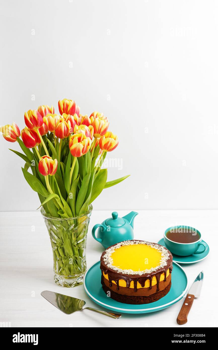Bouquet of tulips, homemade chocolate and orange cheesecake sprinkled with almond slices and cup of tea on white wooden table. Copyspace. Stock Photo