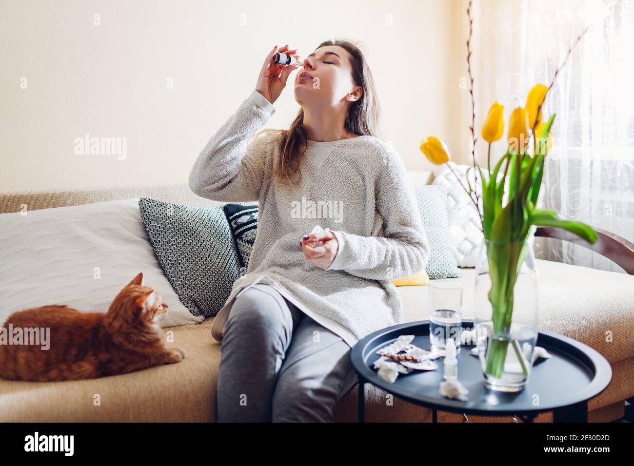 Spring allergy. Woman using nasal drops against seasonal allergy and taking pills at home sitting on couch with cat. Healthcare and medicine Stock Photo