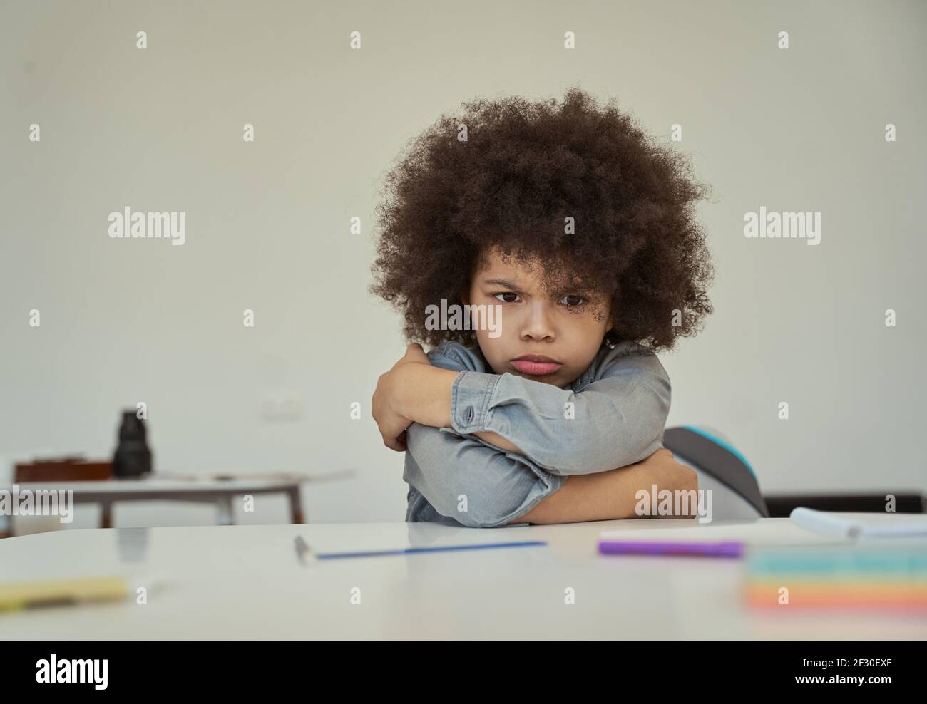 Disgruntled little schoolboy with afro hair looking resentful, frowning while sitting with arms crossed at the table in elementary school classroom Stock Photo