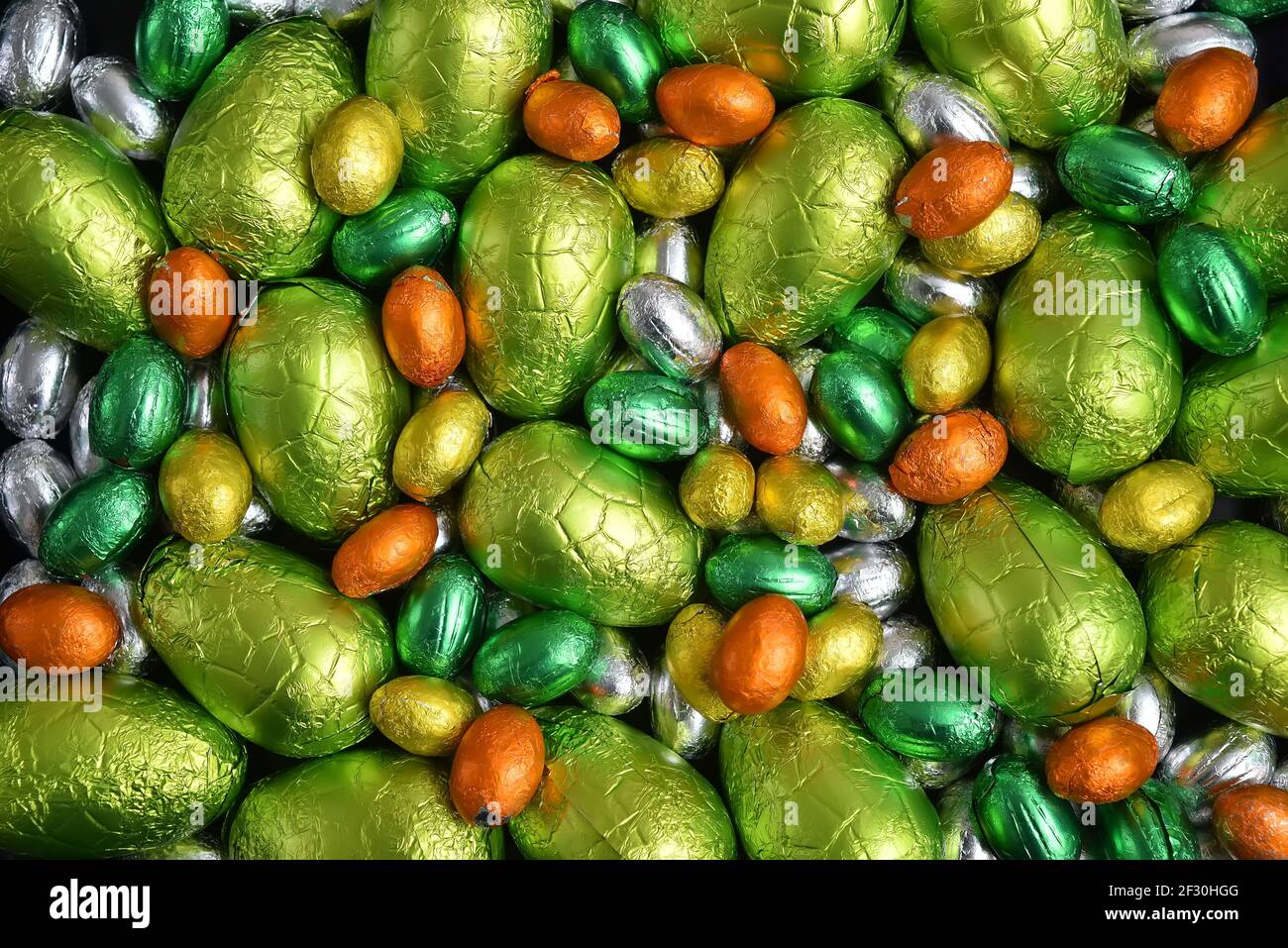 Large & small yellow, gold, orange, green and silver spring colours of foil wrapped chocolate easter eggs, against a black background. Stock Photo