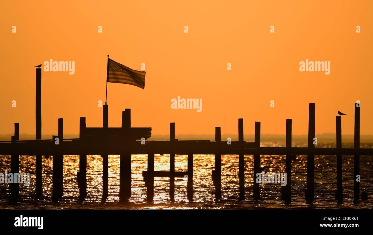 A U.S. flag flies on a pier as the sun sets over Mobile Bay in Fairhope, AL, USA, on Oct. 14, 2020. Stock Photo
