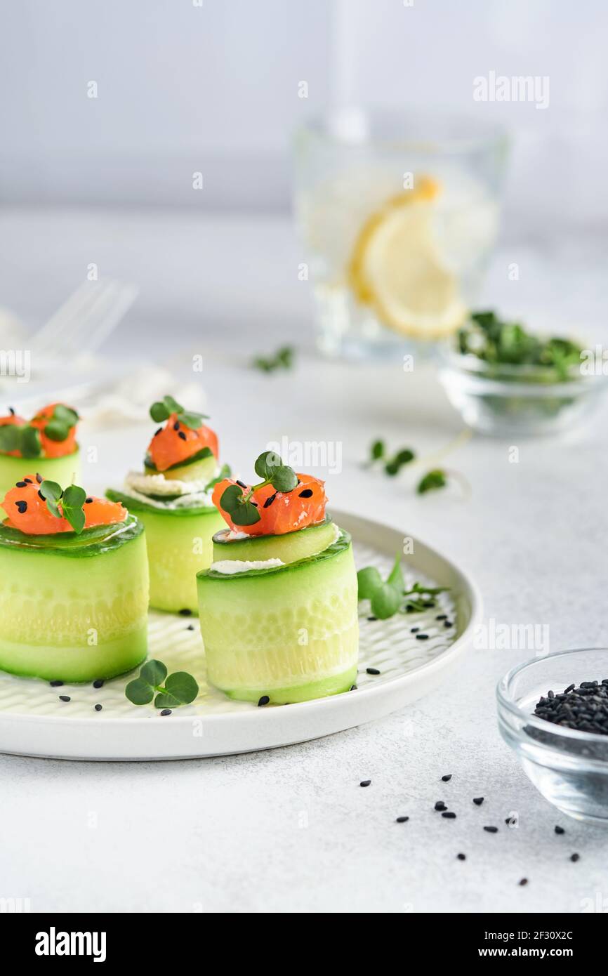 Cucumbers rolls with soft cheese, pieces of salted salmon, microgreens and black sesame served on a white plate. Holiday vegetable appetizers. Selecti Stock Photo