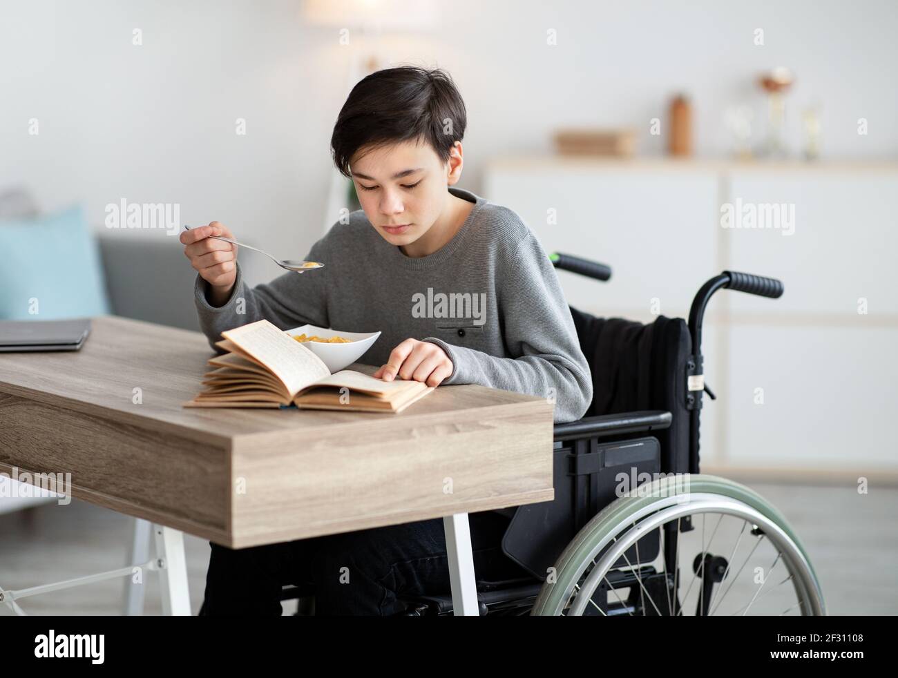 Positive impaired teenager in wheelchair having breakfast and reading book at home. Stay home pastimes Stock Photo