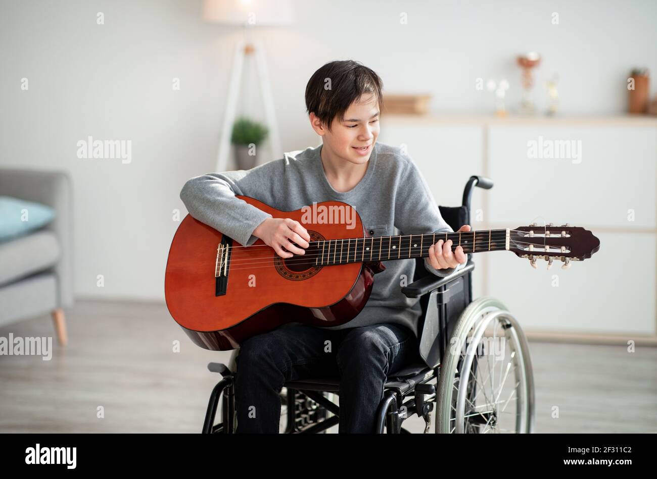 Handicapped teen boy in wheelchair playing guitar and singing indoors. Stay home hobbies Stock Photo