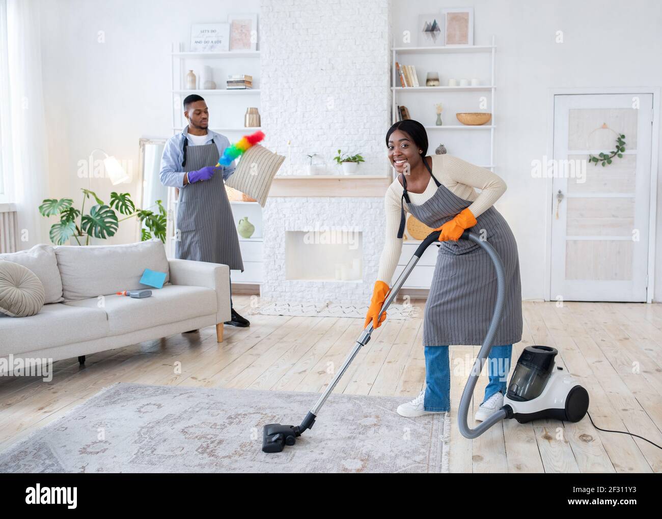 Happy black woman vacuuming floor while her boyfriend dusting cushion