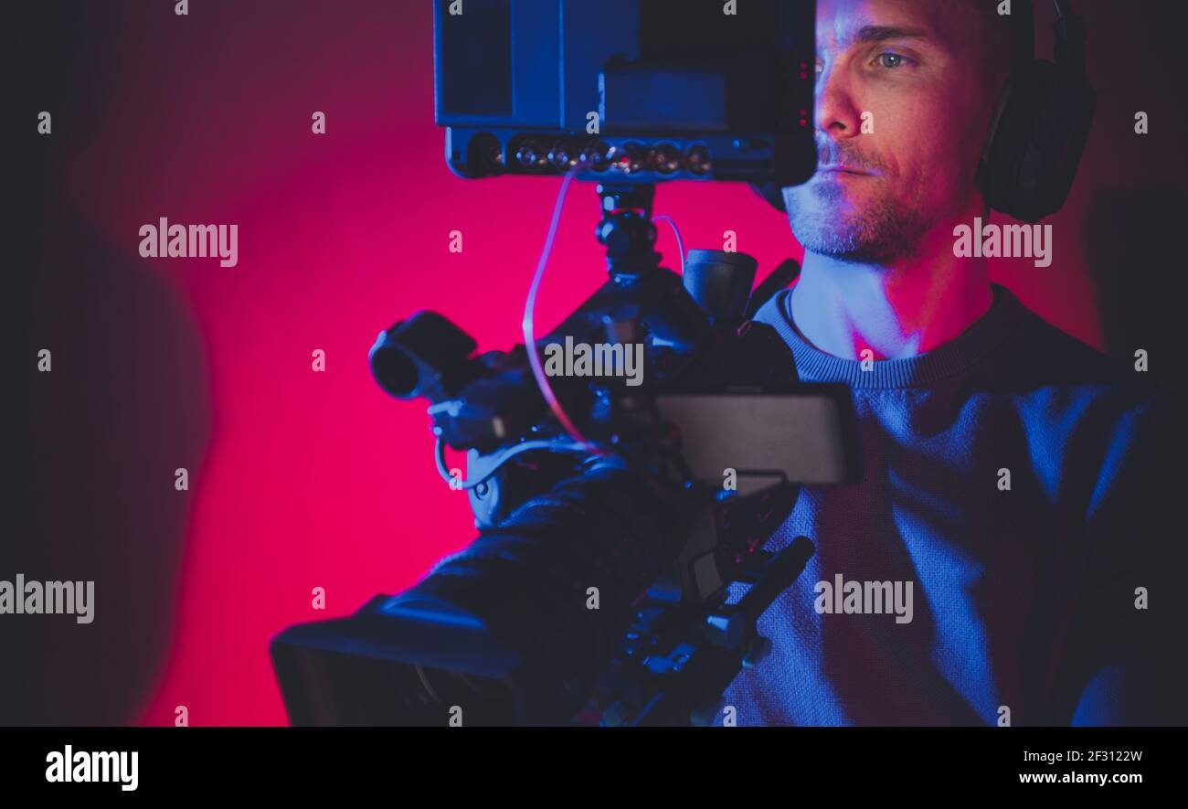 Video Production Stage Dark Red and Blue Illuminated and Film Making Professional Digital Camera Operator at Work. Caucasian Men in His 40s. Stock Photo