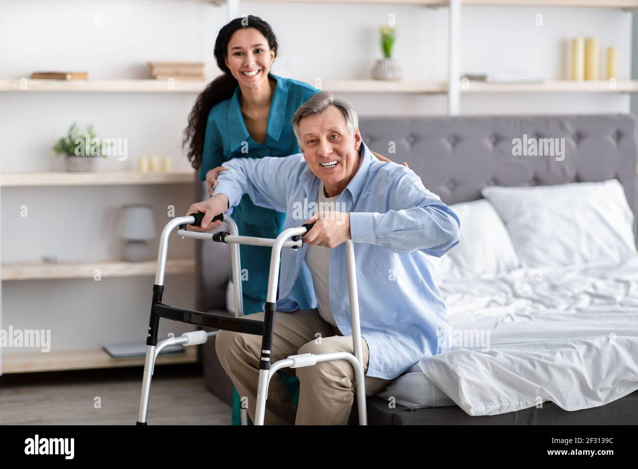 Female nurse helping elderly male with walking frame stand up from bed at home. Professional care for disabled patients Stock Photo