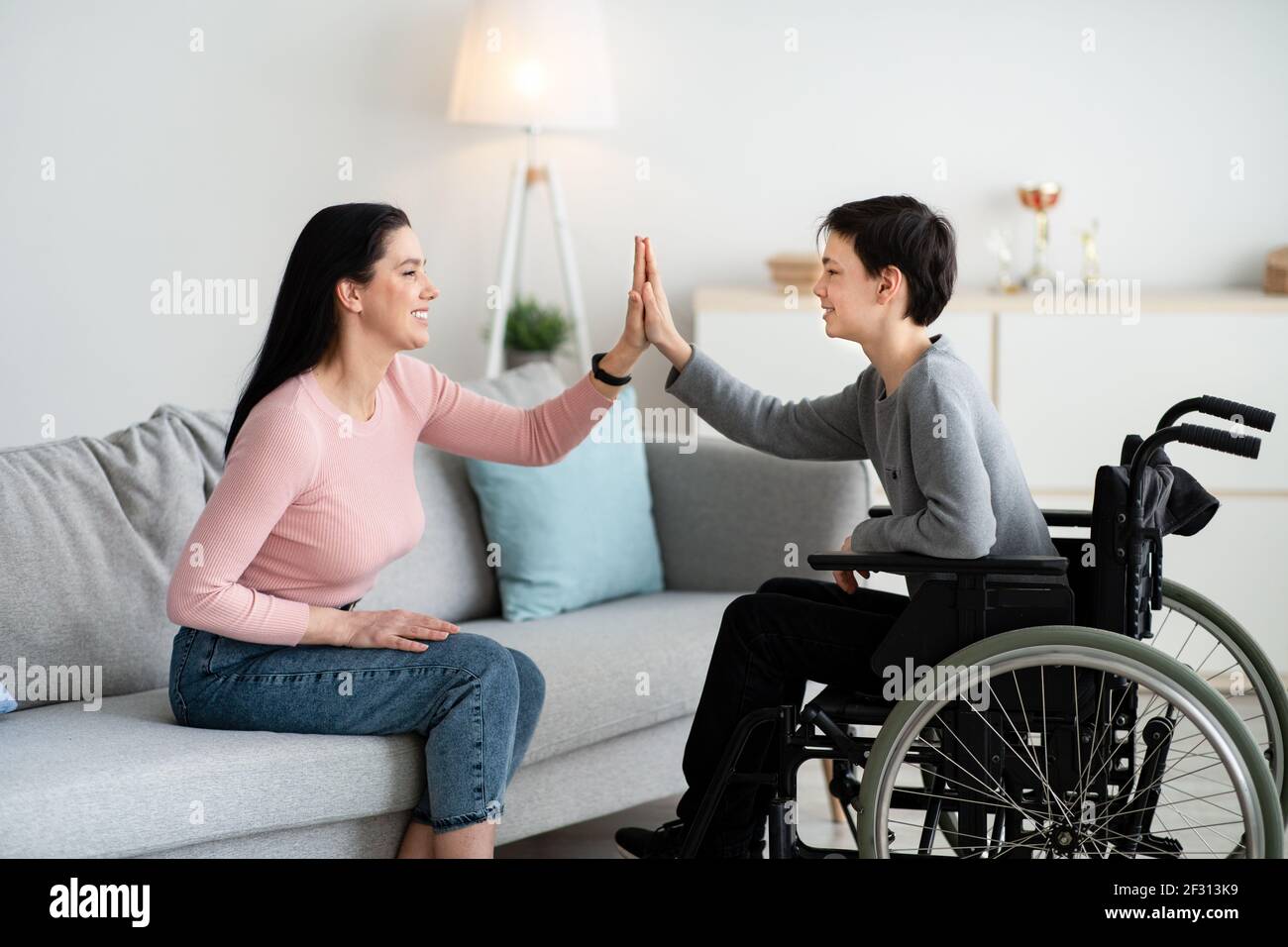 Happy teenage boy in wheelchair high fiving his mother at home, side view Stock Photo