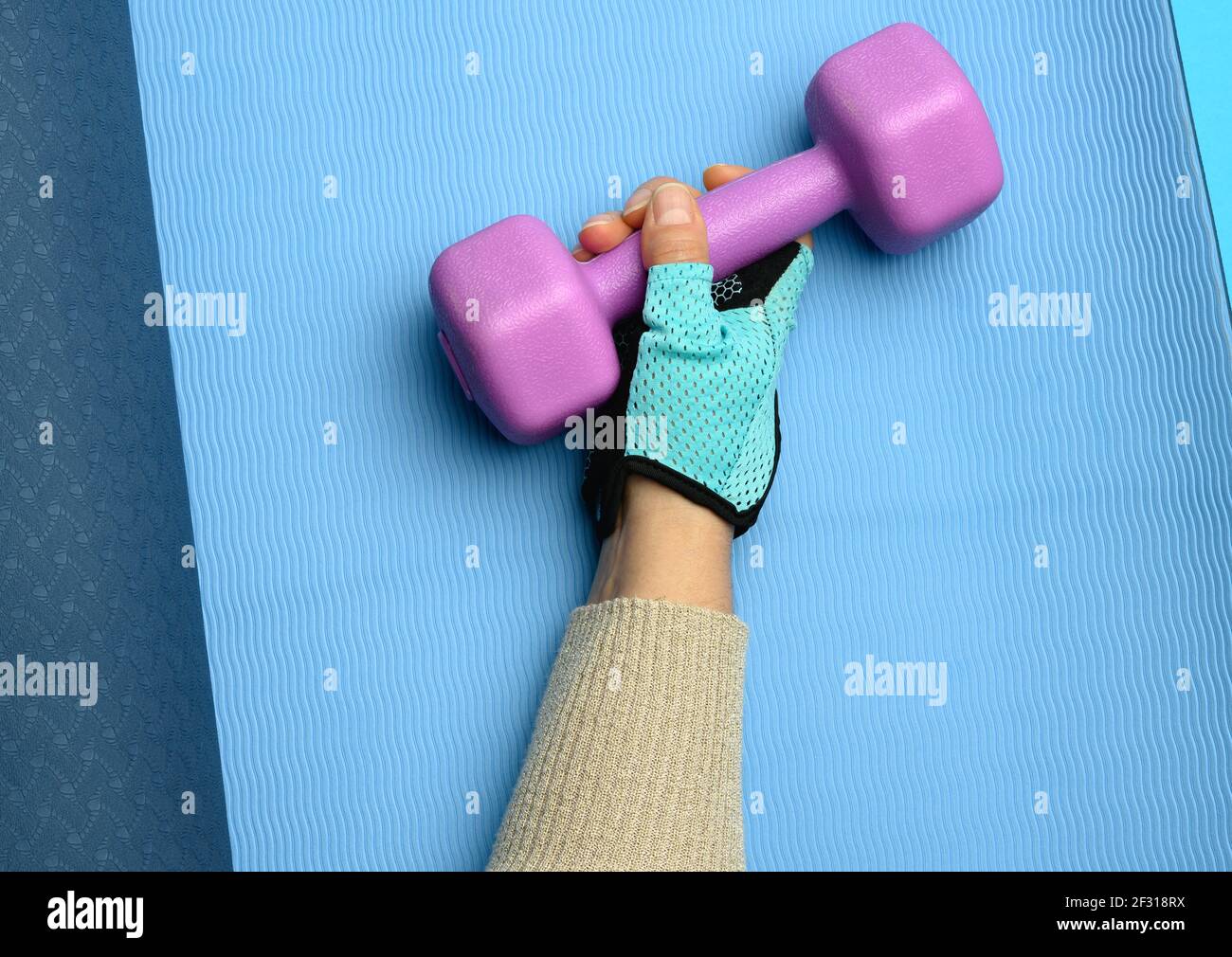 Female hand in a blue sports glove holds a purple one kilogram dumbbell on a blue background, Stock Photo