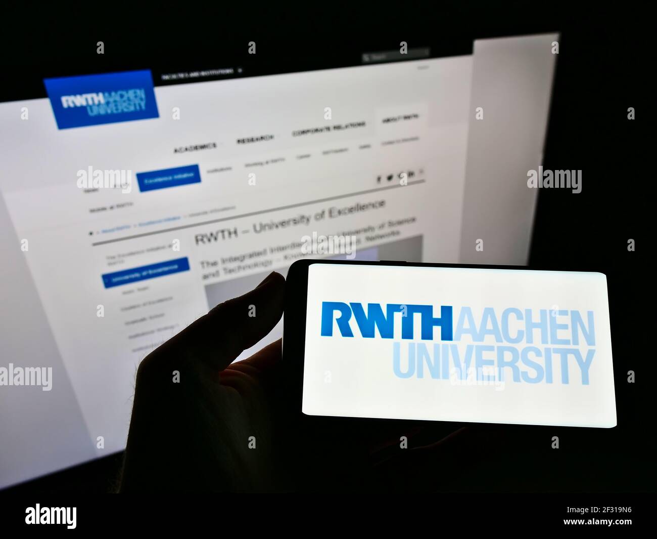 Person holding cellphone with logo of Rheinisch-Westfälische Technische Hochschule Aachen on screen in front of web page. Focus on phone display. Stock Photo