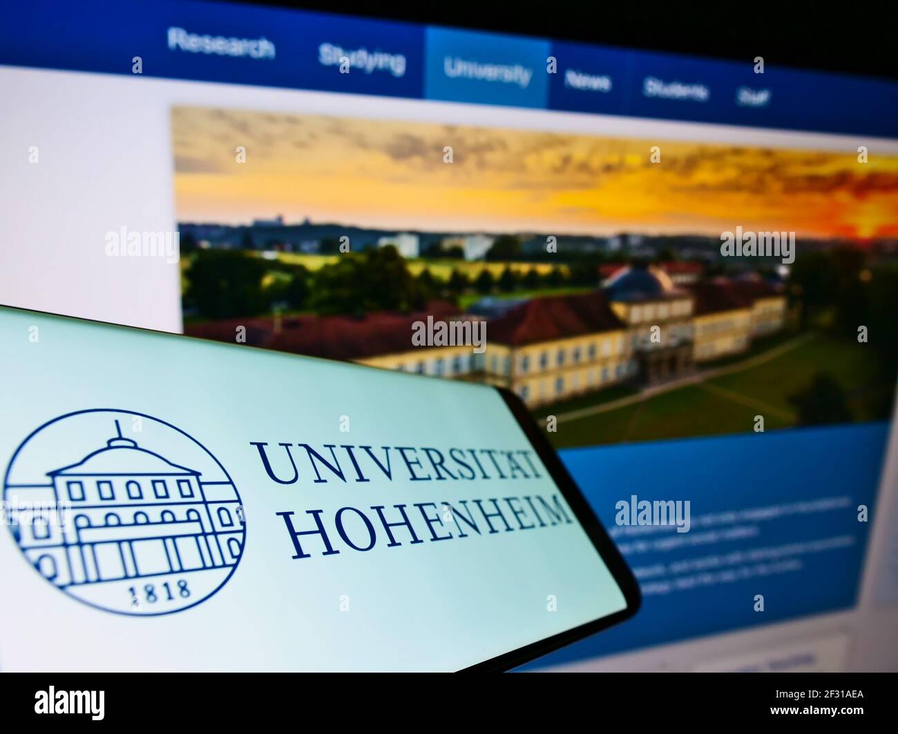 Smartphone with logo of German education institution Universität Hohenheim on screen in front of web page. Focus on center-left of phone display. Stock Photo