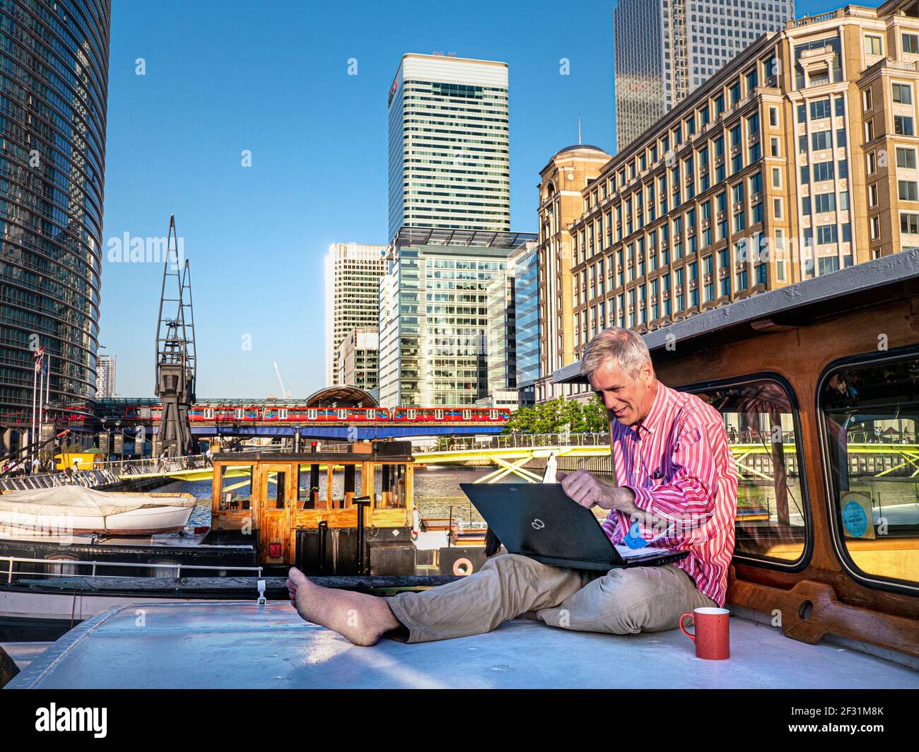 Home working mature man 50-55 outside on his houseboat barge, working on his computer contrasted with the surroundings of office buildings, hotels and cross rail tube train in background. Canary Wharf London UK.  Telecommuting, also called remote working, future of work, telework, teleworking, working from home, mobile work, remote job, work from anywhere, and flexible workplace, a work arrangement in which employees do not commute or travel to a central place of work, such as an office building, warehouse, or store. Stock Photo