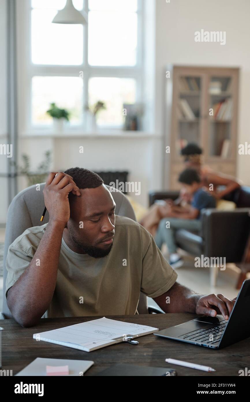 Pensive young African man with pencil in hand sitting by table in front of laptop and scrolling through online data against his family Stock Photo