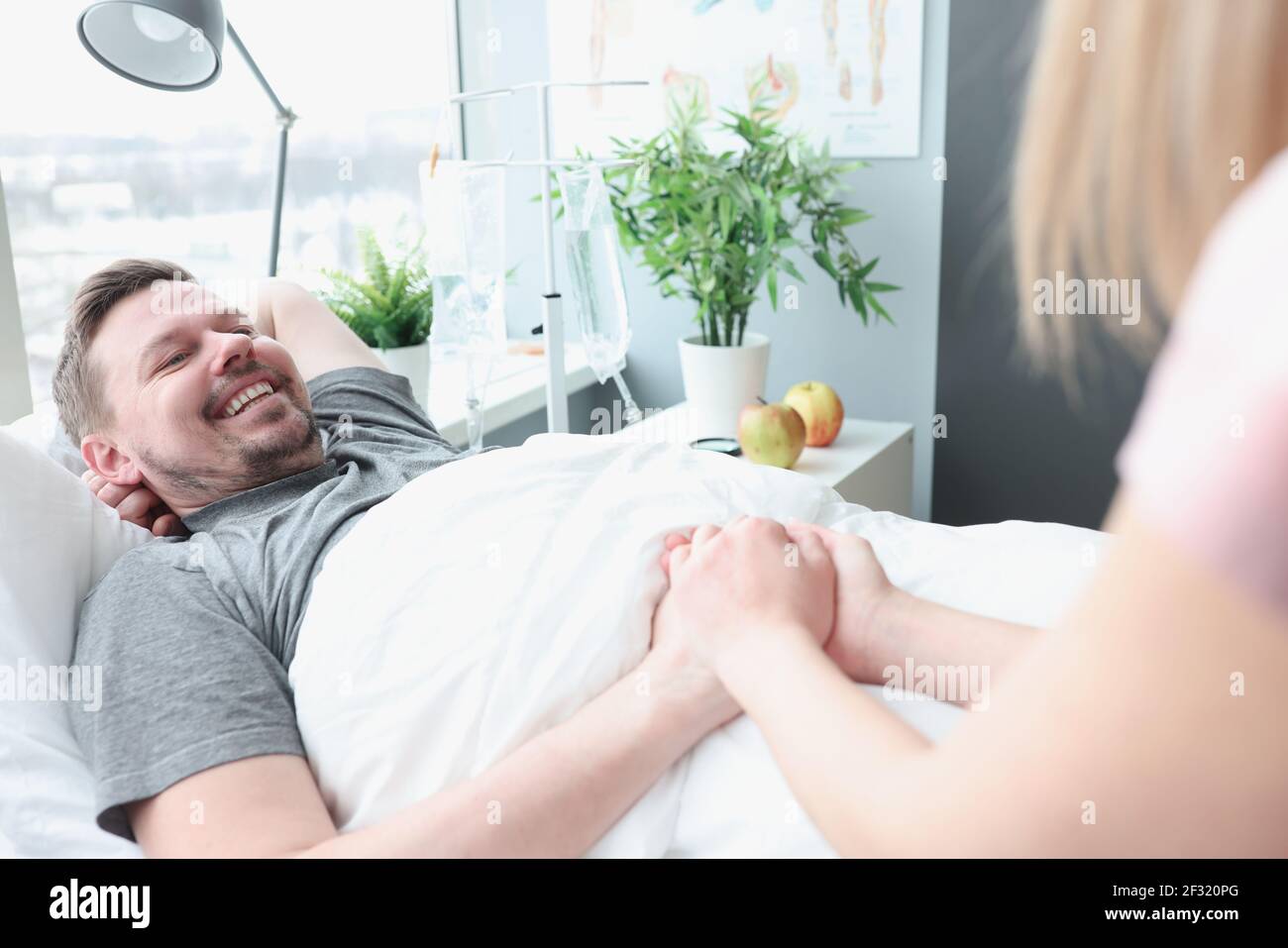 Smiling man lies on hospital bed his hand is held by woman Stock Photo