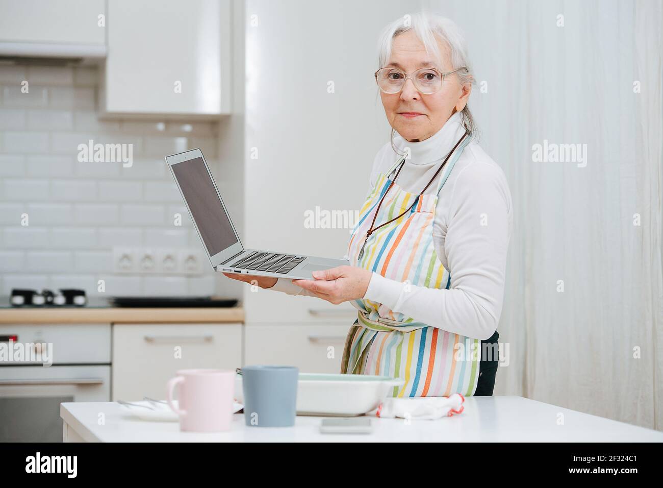 Portrait of a granny talking via webcam on her laptop in the kitchen,  sharing her progress. She's looking at the camera Stock Photo - Alamy