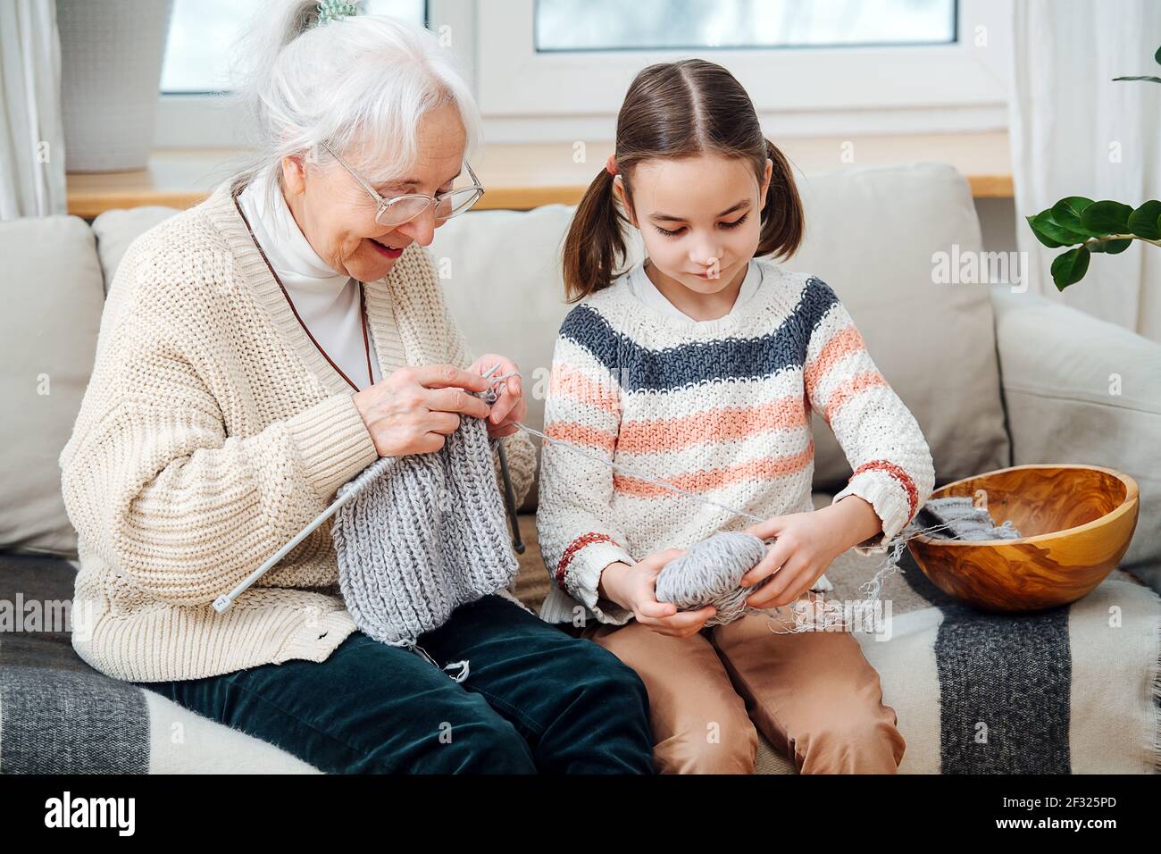 Cheerful granny knitting on a couch at home, her granddaughter helping her by unwinding ball of yarn. Stock Photo