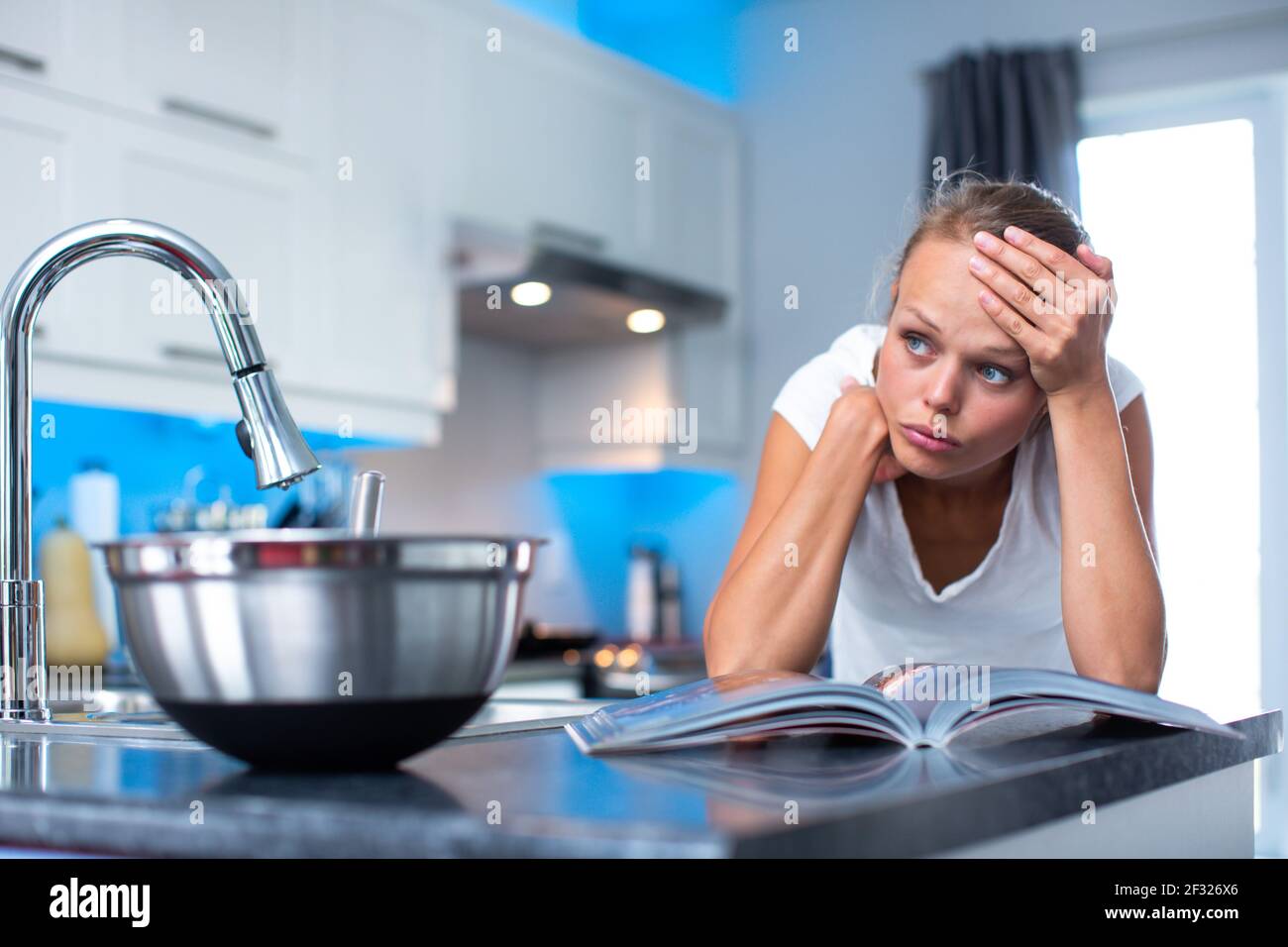Pretty, young woman in her modern, clean and bright kitchen, thinking ...