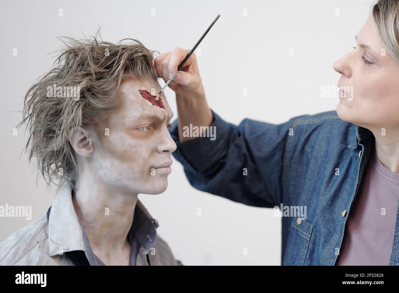 Contemporary visage artist with brush applying zombie makeup on face of young businessman or actor against white wall in office or studio Stock Photo