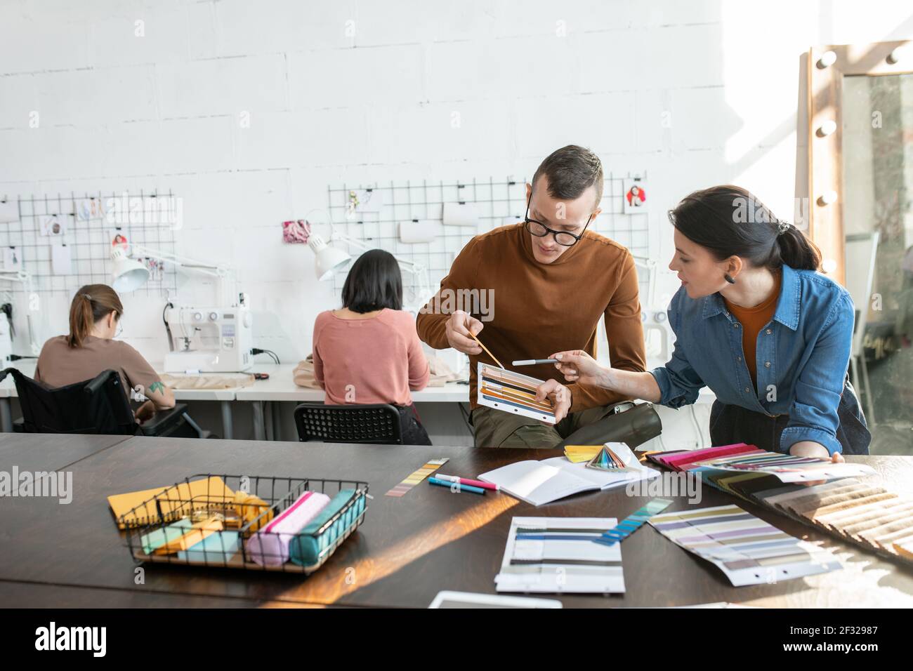 Young contemporary fashion designers choosing colors and textile for new collection against two females using sewing machines in workshop Stock Photo