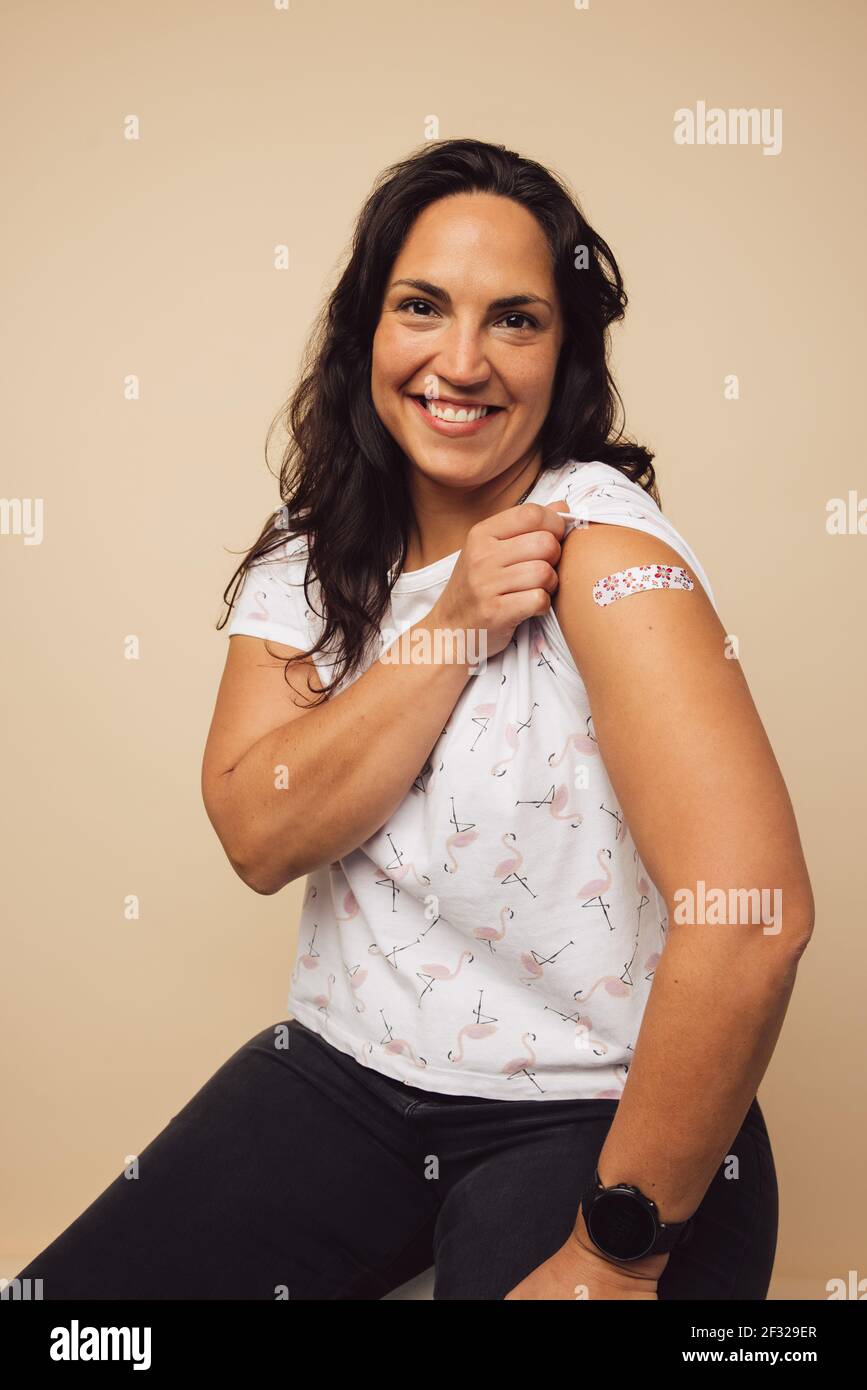 Happy mid adult woman showing a bandage on her arm after she received vaccination. Woman feeling proud to get inoculated. Stock Photo