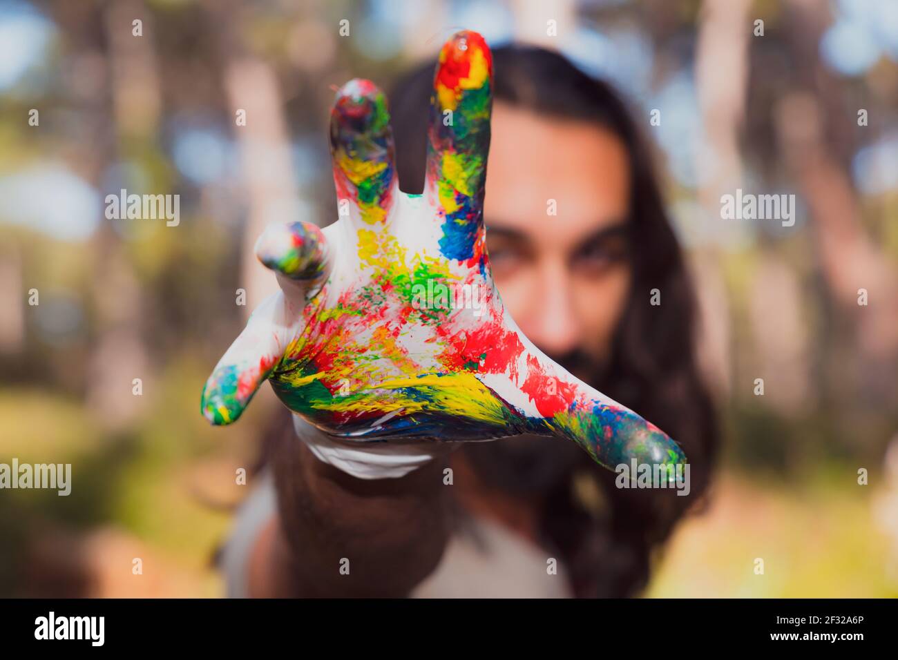 Young brunette male with long hair shows his palm, hand painted in different colors, Outdoor body paint activity, Holi Festival at Park. People. Stock Photo