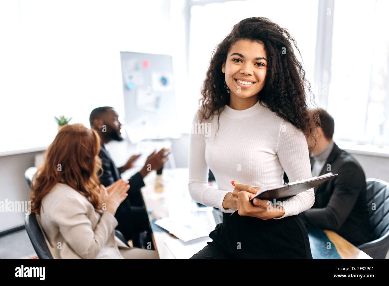 Portrait of beautiful young business woman at the briefing meeting. Successful african american female in formal wear looks directly at the camera, smiling, work together with colleagues Stock Photo