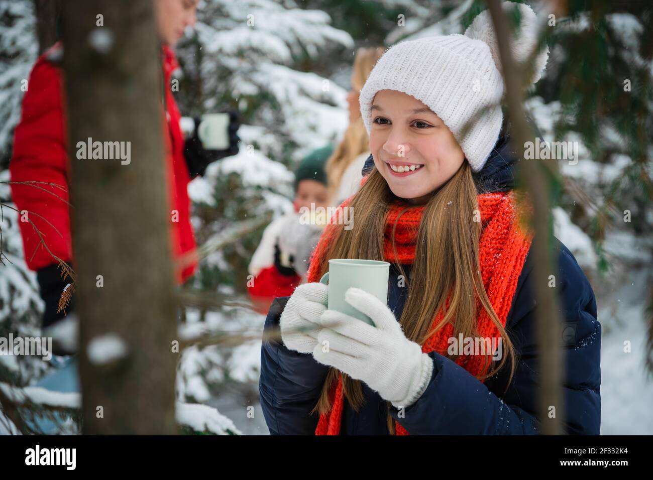 A smiling girl drinks warm coffee with her family in a beautiful snow-covered forest before the new year. Active lifestyle and walking in the fresh Stock Photo