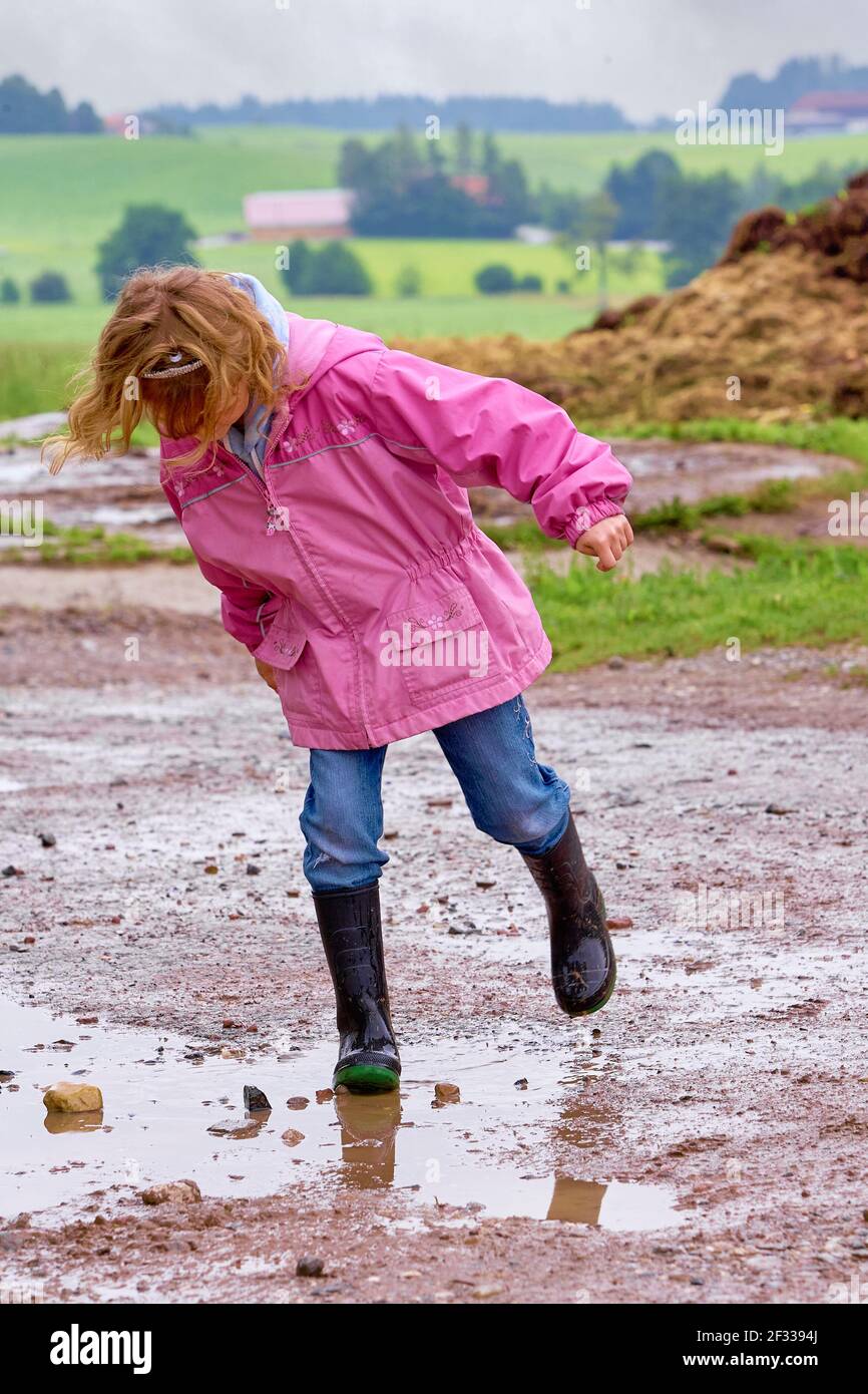 Full body girl in pink raincoat and rubber boots playing in puddle of mud in countryside Stock Photo