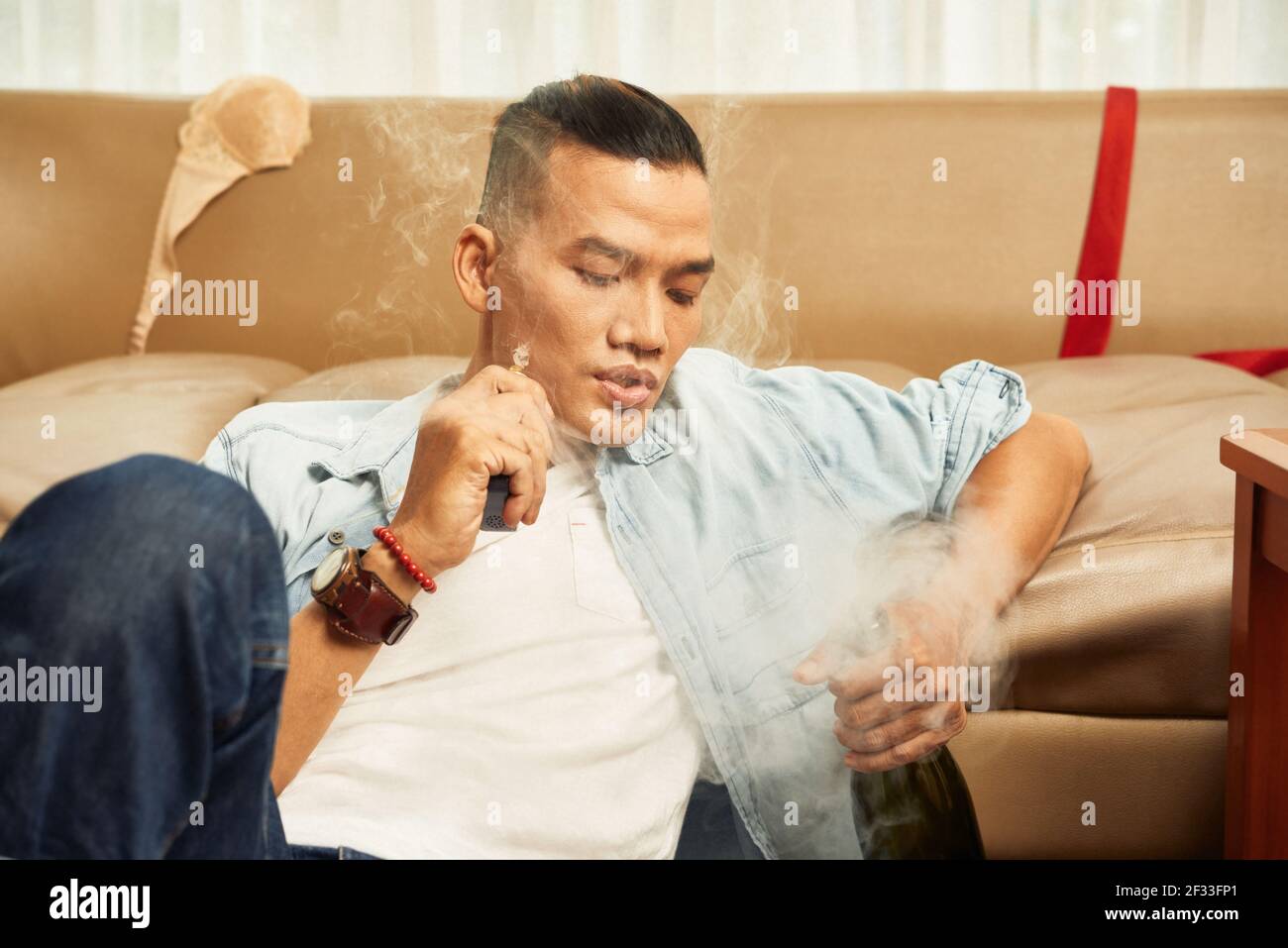 Handsome young Asian man smoking cigarette and drinking alcohol at home Stock Photo
