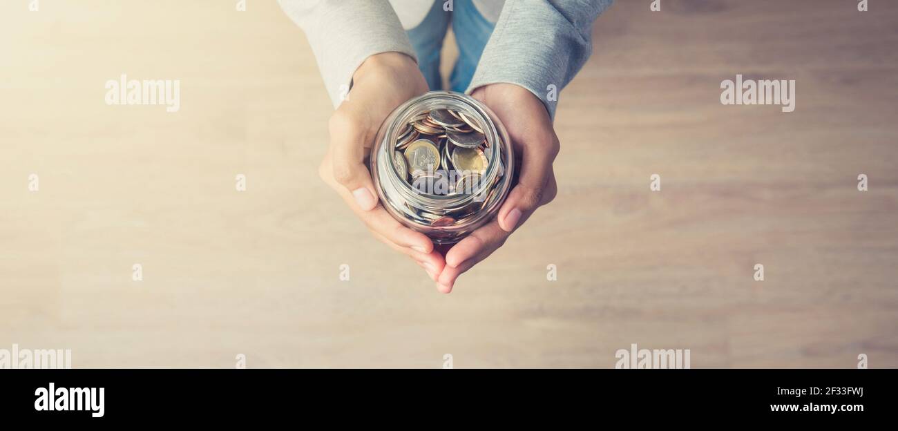 Young woman hands holding glass jar with money (coins) inside - panoramic banner background, top view, with copy space Stock Photo