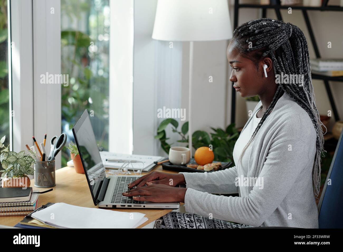 Serious young Black woman with dreadlocks working on laptop at her desk at home and writing essay for colleage Stock Photo