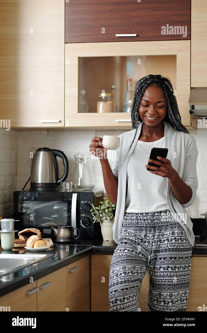 Cheerful young Black woman in loungewear drinking cup of morning coffee in  her kitchen and laughing at funny memes on smartphone screen Stock Photo -  Alamy