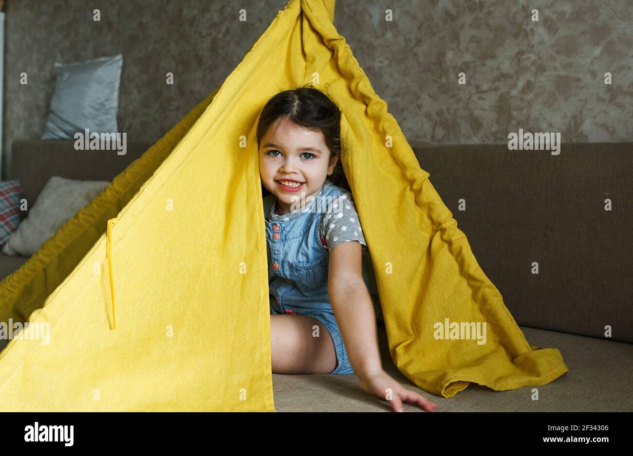 little girl playing at home on the couch hiding in a yellow teepee ...