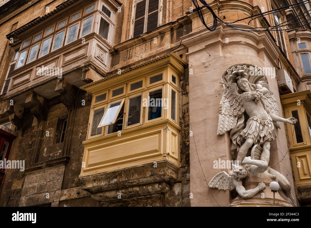 Traditional Maltese houses with wooden covered balconies and St Michael the Archangel vanquishing the devil statue on building corner in city of Valle Stock Photo