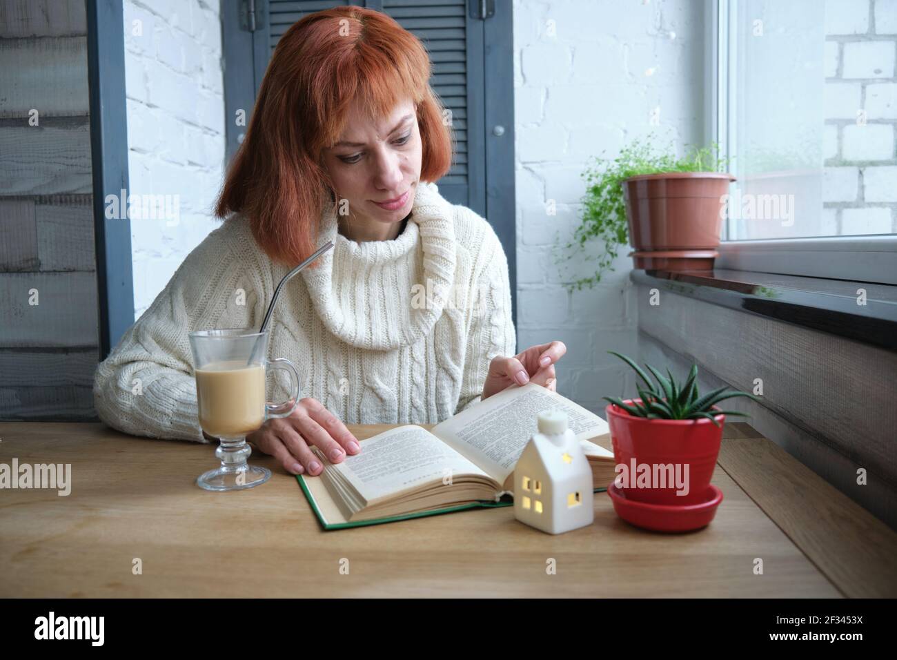 Young adult red haired girl relaxing at home with mug of hot beverage and book in domestic kitchen, relaxing during quarantine Stock Photo