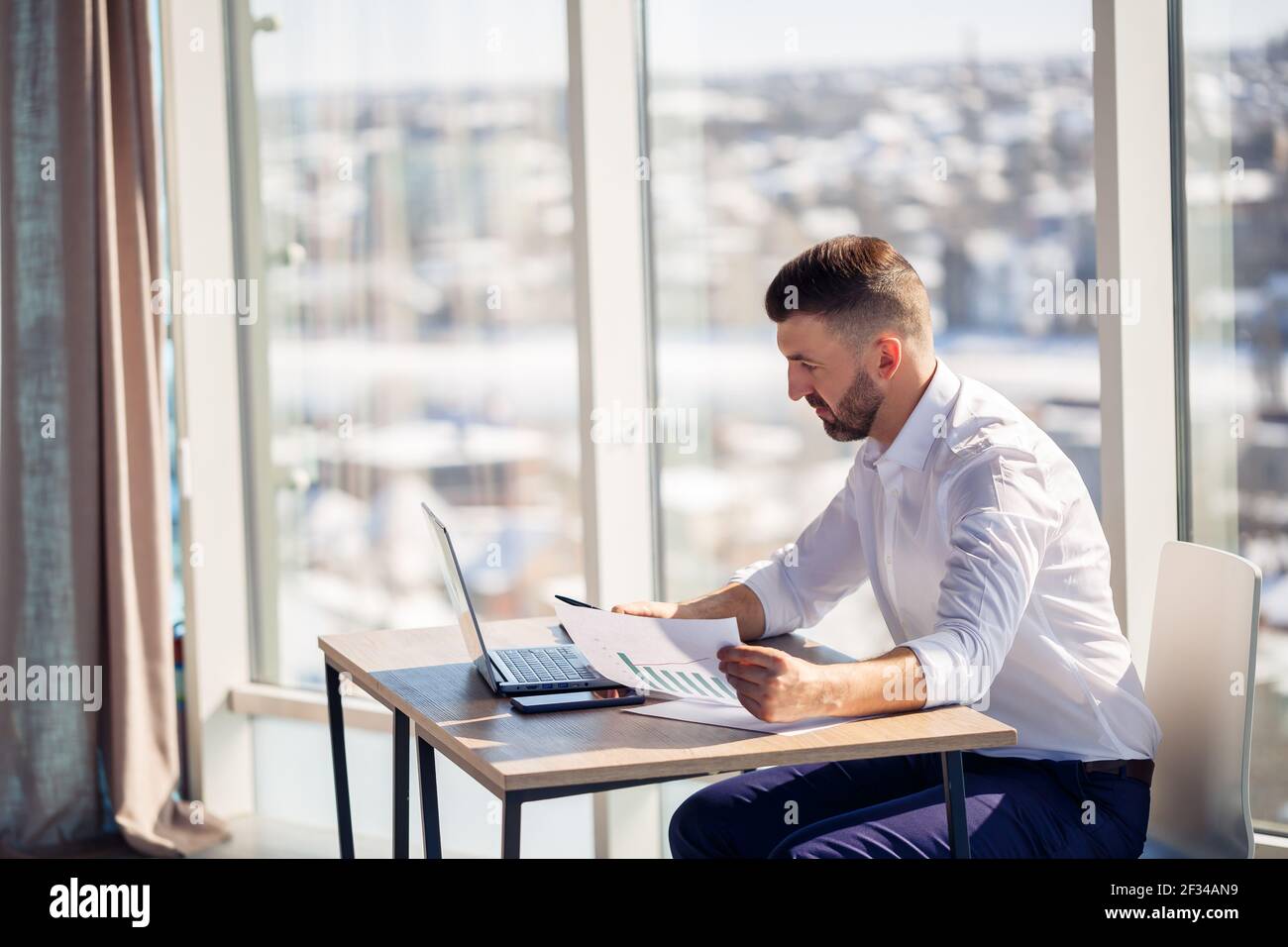 An adult successful male businessman sits in his office with large windows and works at a laptop, working on new projects. Stock Photo