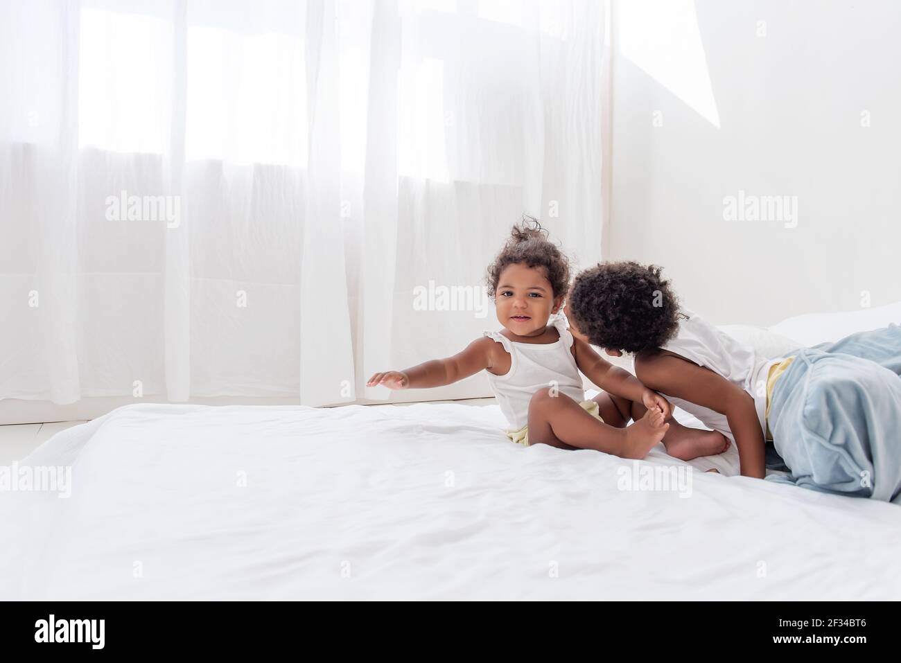 Brother and sister African Americans play together on white bed in loft interior. Siblings having fun among the blue pillows in the morning. Boy kissi Stock Photo