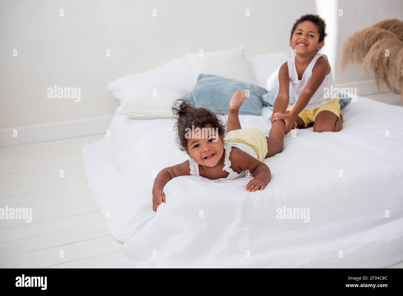 Brother and sister African Americans play together on white bed in a loft interior. Siblings having fun among the blue pillows in the morning. Boy pul Stock Photo